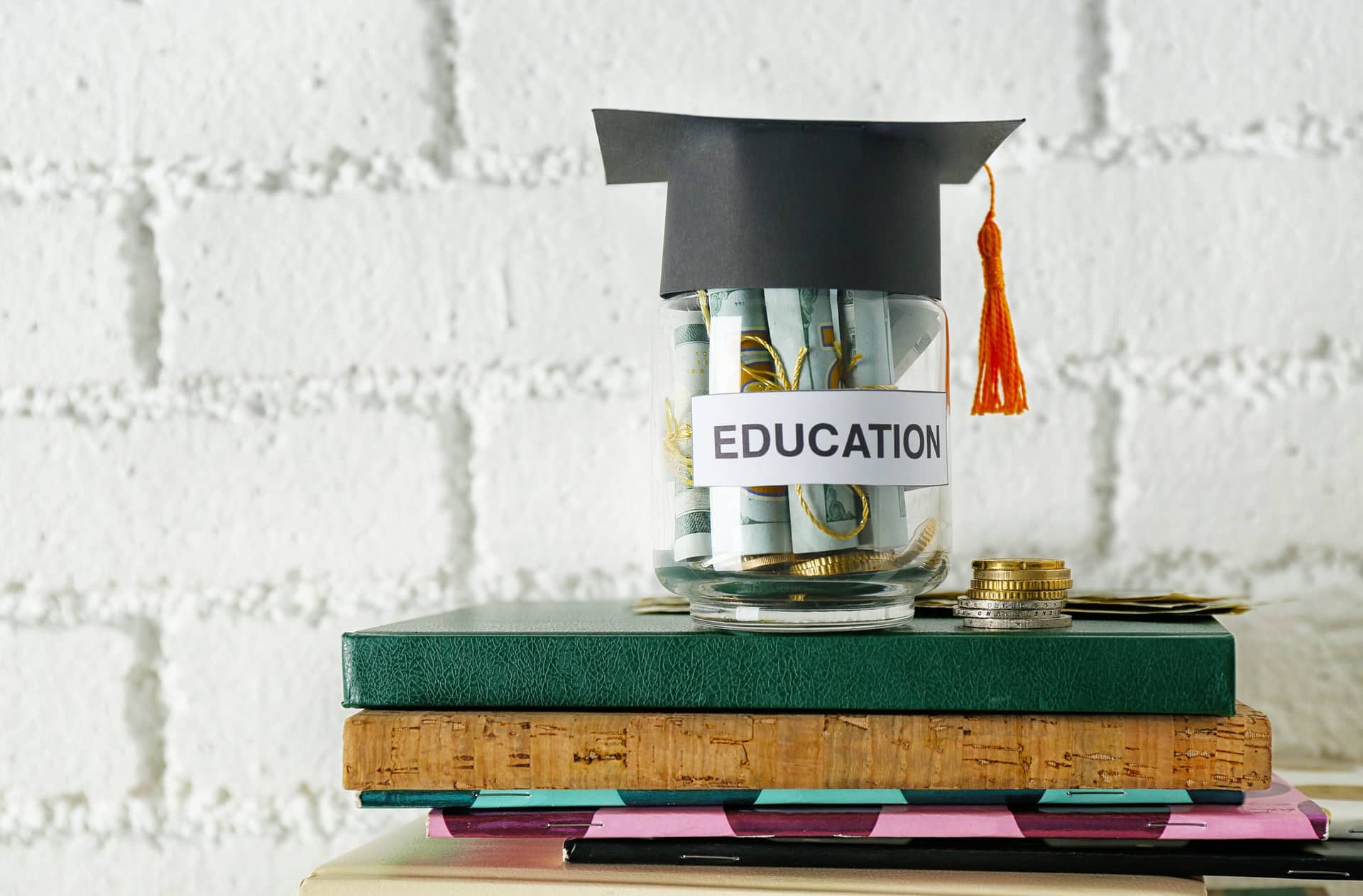 Glass jar with money for education on pile of books and notepads against brick wall